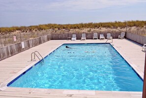 A rectangular swimming pool with a wooden deck, surrounded by a fence and beach vegetation. - Cute, spacious 1-bedroom oceanfront condo with free WiFi, an outdoor pool, and an expansive balcony located midtown! (Ocean City)