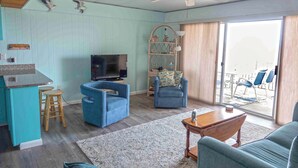 A living room with a TV, blue chairs, a wooden coffee table, and a view through a sliding glass door. - Cute, spacious 1-bedroom oceanfront condo with free WiFi, an outdoor pool, and an expansive balcony located midtown! (Ocean City)
