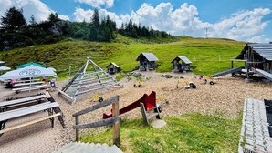 Un espace extérieur situé en zone rurale, aménagé avec des tables de pique-nique, une aire de jeux et de petites structures en bois.