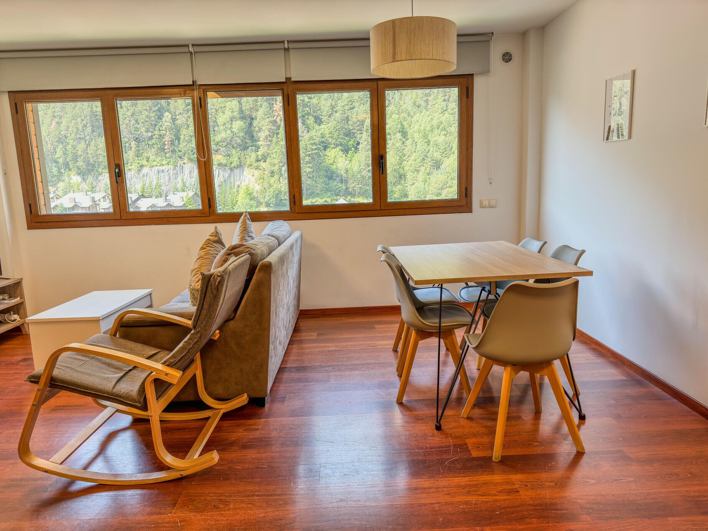 Una habitación con una mesa de comedor de madera, sillas, una mecedora, un sofá y un amplio ventanal con vista al bosque.