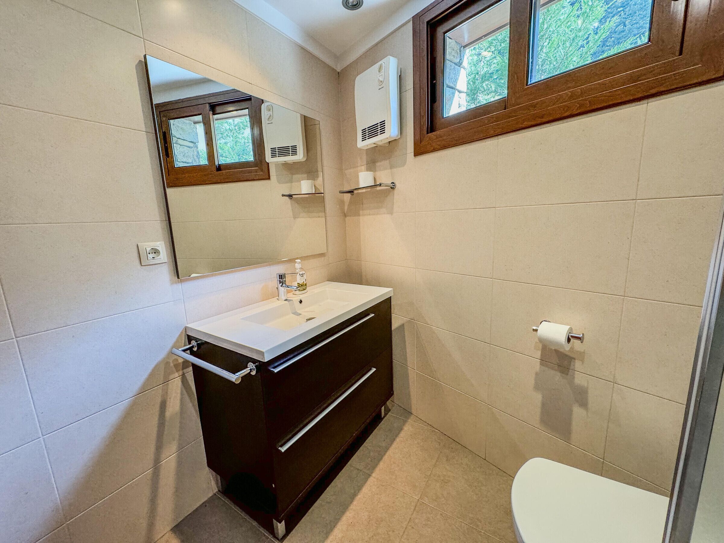 A bathroom with a white sink, dark cabinetry, a mirror, a window, and a toilet.