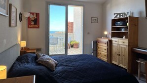 A bedroom with a bed, a desk, a wardrobe, a lamp, a poster, and a view of the outside. - Studio sea view, parking - 2 guests (Banyuls-sur-Mer)