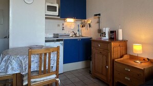 A compact kitchen with a microwave, blue cabinets, a wooden table with a fish-patterned tablecloth, and a bedside lamp. - Studio sea view, parking - 2 guests (Banyuls-sur-Mer)