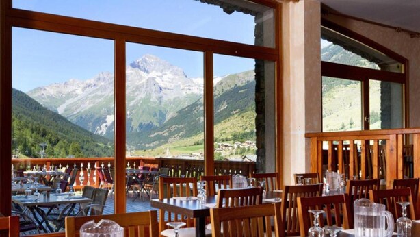 Un restaurant avec des tables dressées pour le service, de grandes fenêtres offrant une vue sur les montagnes et une terrasse en bois.