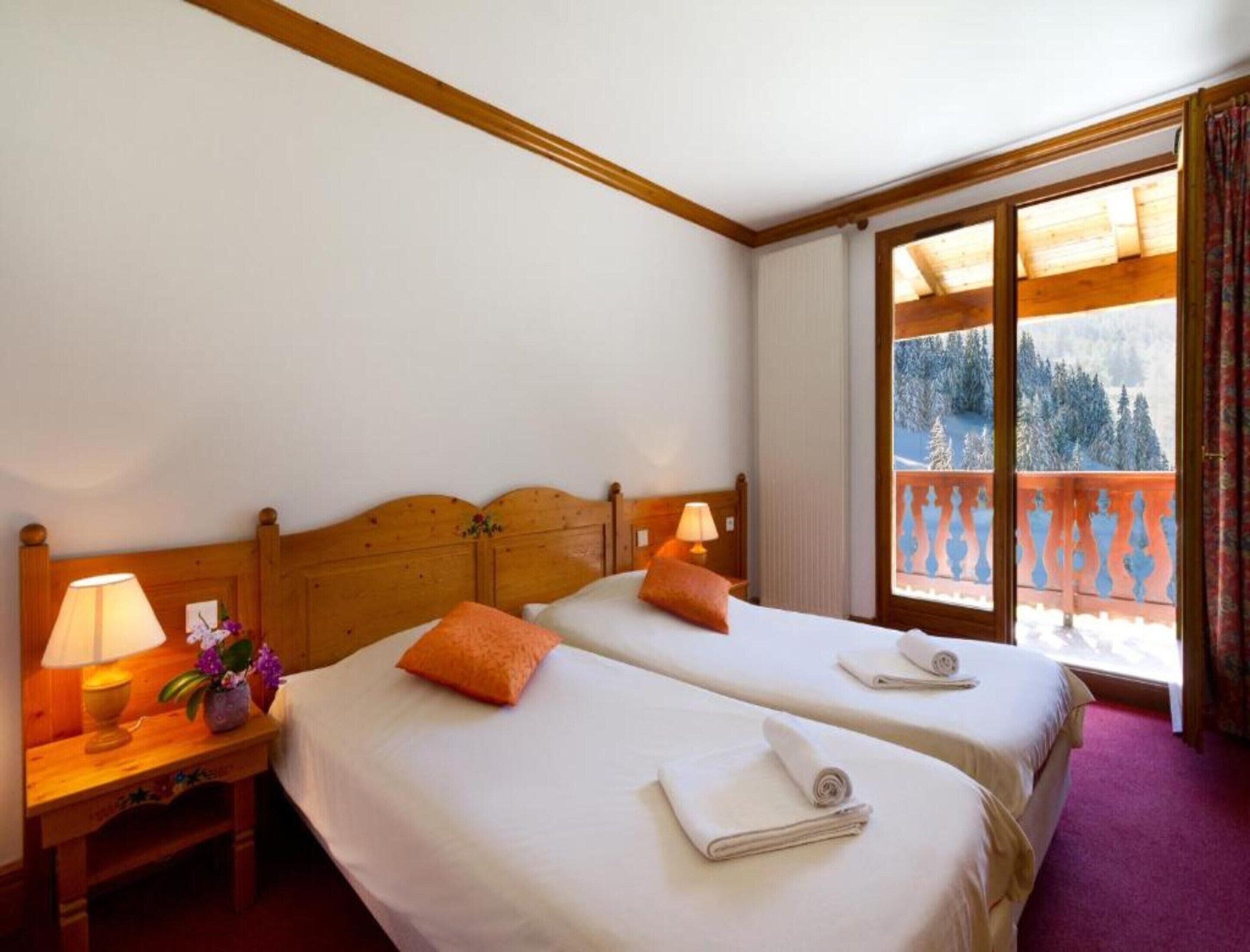 A hotel room with two beds, a wooden headboard, a nightstand with a lamp, and a window with a view of a snowy landscape.