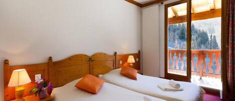A hotel room with two beds, a wooden headboard, a nightstand with a lamp, and a window with a view of a snowy landscape.