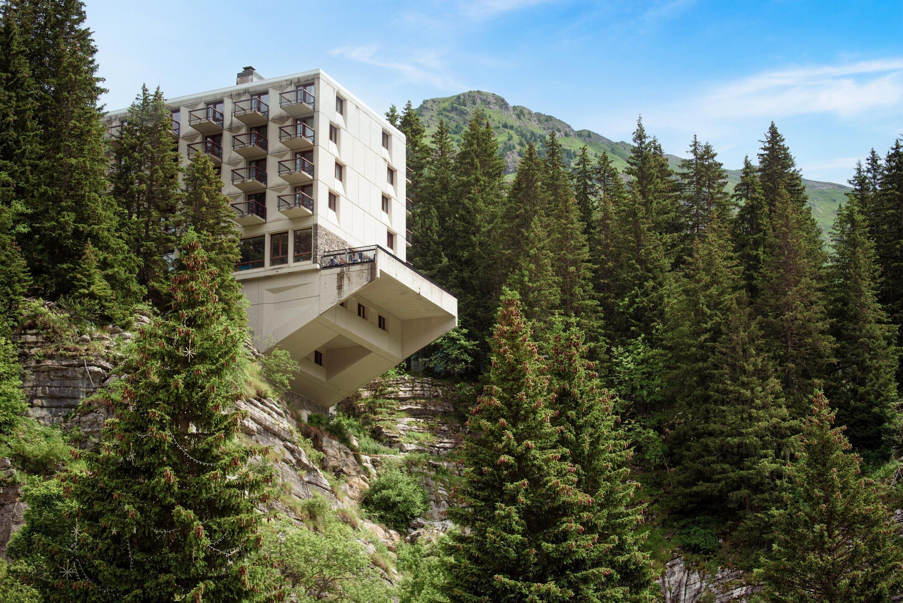 A modern building with balconies, surrounded by dense forest and mountains.
