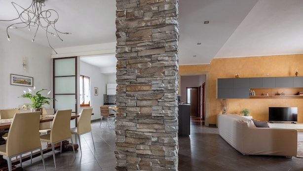 Una sala moderna con un pilar de piedra, un comedor con sillas y una mesa, una cocina con armarios y un televisor.