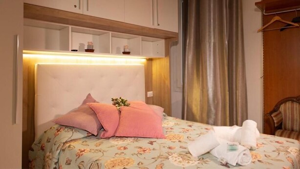 Une chambre d’hôtel avec un lit, des oreillers, une tête de lit, une étagère en bois, un rideau et une chaise.