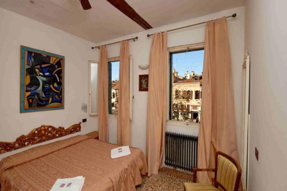 Een kamer met een bed, een stoel, een radiator, een raam met gordijnen en een schilderij aan de muur.