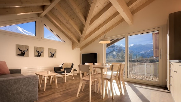 Uma sala de estar ampla com vigas de madeira, sofá, mesa de jantar, cadeiras, obras de arte e uma varanda com vista para as montanhas.