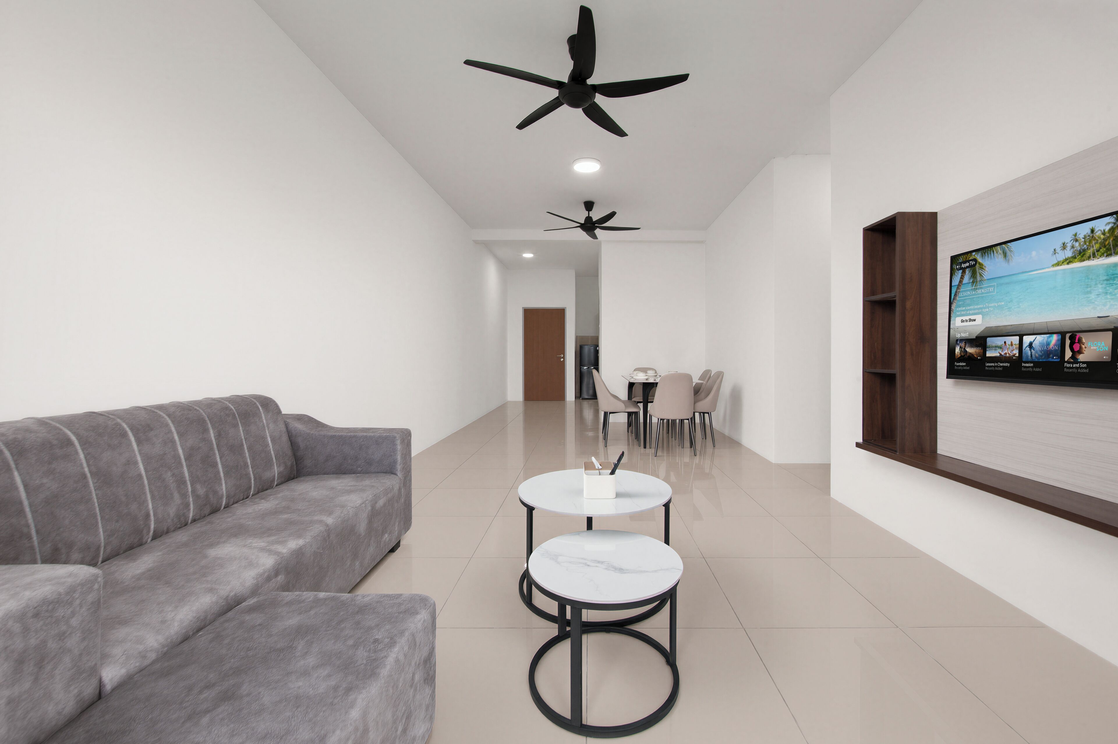 Un salon moderne avec un canapé gris, deux tables rondes, des ventilateurs de plafond, un téléviseur et un coin repas.