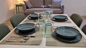 Uma área de refeições com mesa posta para quatro pessoas, um sofá, uma luminária e uma janela com vista para o verde.