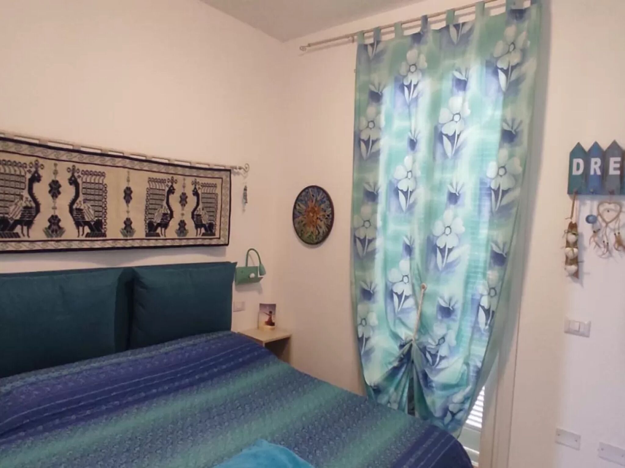 Een slaapkamer met een bed, een hoofdeinde, een nachtkastje, een wandklok, een gordijn en een decoratie aan de muur.