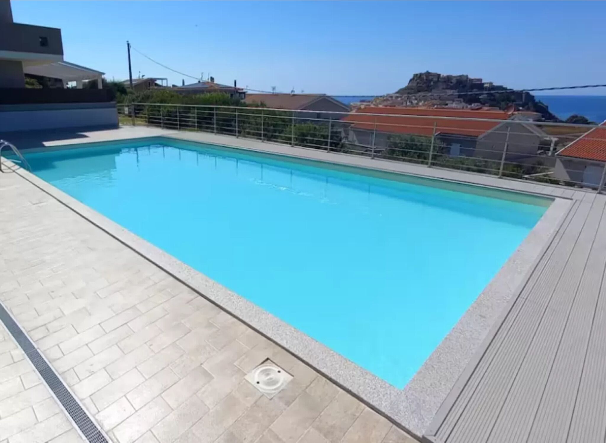 Una piscina rectangular con superficie de agua azul cristalina, rodeada por un solado de azulejos y una cerca de metal.