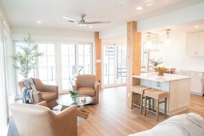 A spacious living area with a glass coffee table, two armchairs, a sofa, a kitchen island with stools, and a ceiling fan.