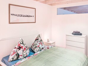 Ein Schlafzimmer mit Bett, Kissen, einem Nachttisch, einer Kommode, einem gerahmten Bild und einem Oberlicht.