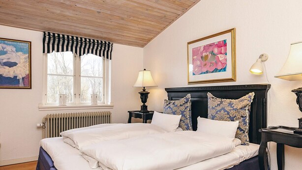Un dormitorio con cama, radiador, ventana con cortinas, mesita de noche con lámpara y dos cuadros en la pared.
