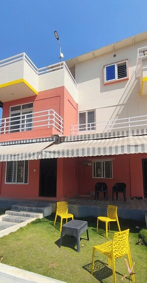 A two-story building with a red and white facade, a covered outdoor seating area with yellow chairs, and a staircase leading to the upper floor. - Nandi Holiday Homestay - Unit 2 (Thirnahalli)