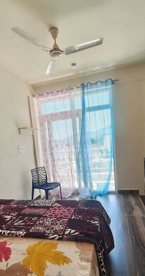 Une chambre à coucher avec un lit, un ventilateur de plafond, une chaise et une fenêtre avec des rideaux légers.