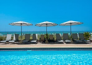 Ein Poolbereich mit Liegestühlen und Sonnenschirmen, mit Blick auf das klare Meer und einem gefliesten Pool.