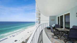 Een balkon met uitzicht op een strand en de oceaan, een eettafel met stoelen, en een bijzettafeltje met een decoratie.