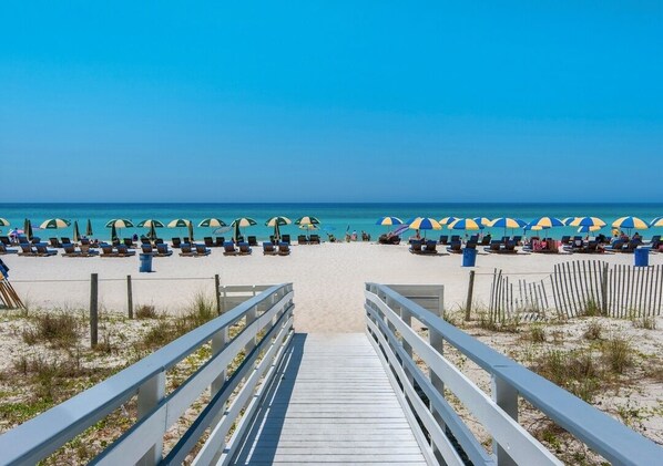 Een strand met een houten looppad naar het zand, rijen strandstoelen en parasols.