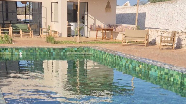 Ein klares Schwimmbecken mit gefliesten Rändern, einer Holzterrasse und Gartenmöbeln.