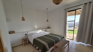 Ein Schlafzimmer mit Bett, Nachttischen, einer Bank, einer Pendelleuchte und Blick durch ein Fenster.