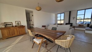 Ein geräumiger Wohnbereich mit Esstisch aus Holz, Korbstühlen, einer Couch und einem großen Fenster mit Aussicht.