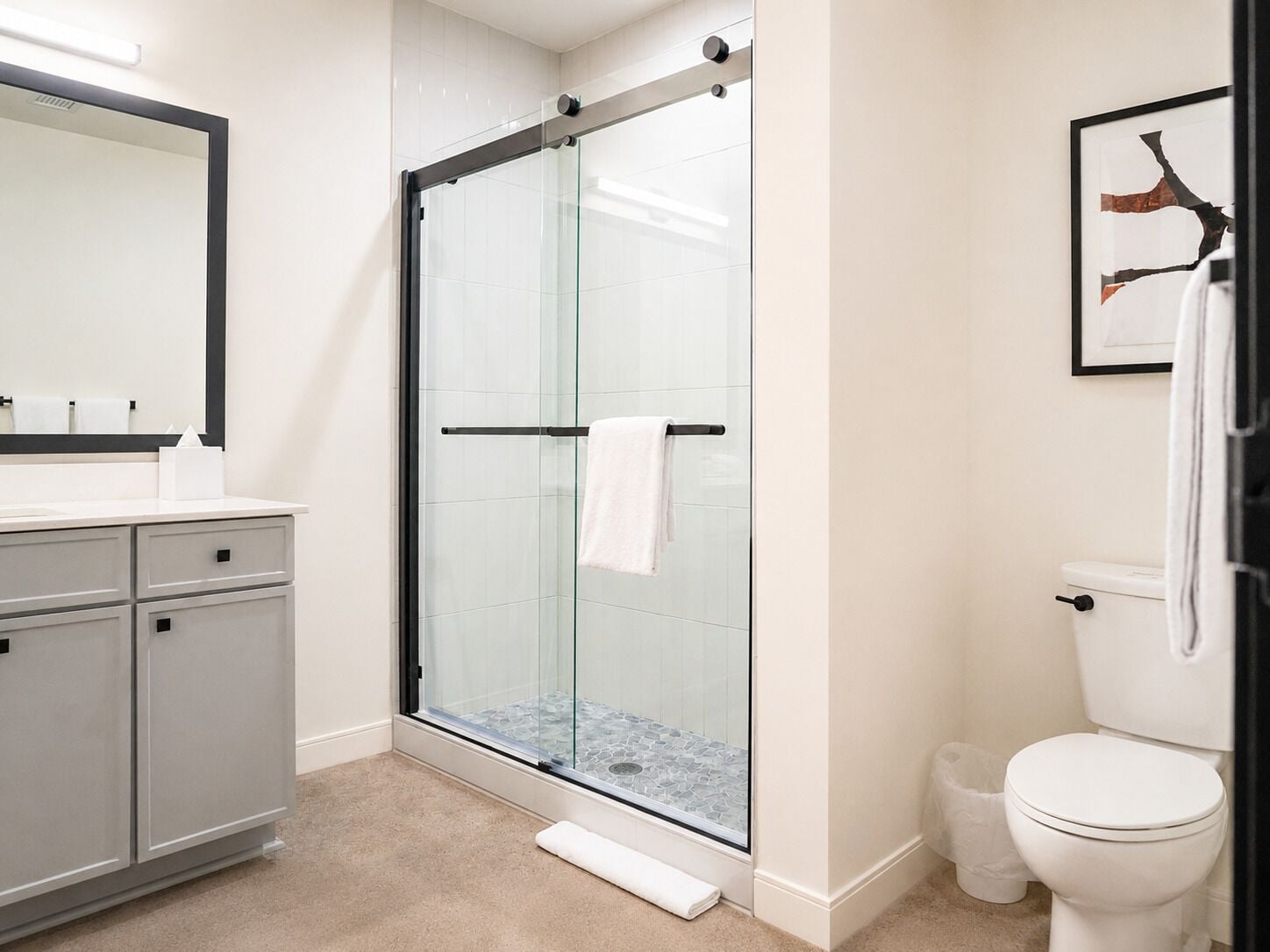 A bathroom with a glass shower enclosure, a white toilet, a sink with a mirror, and a framed picture on the wall.