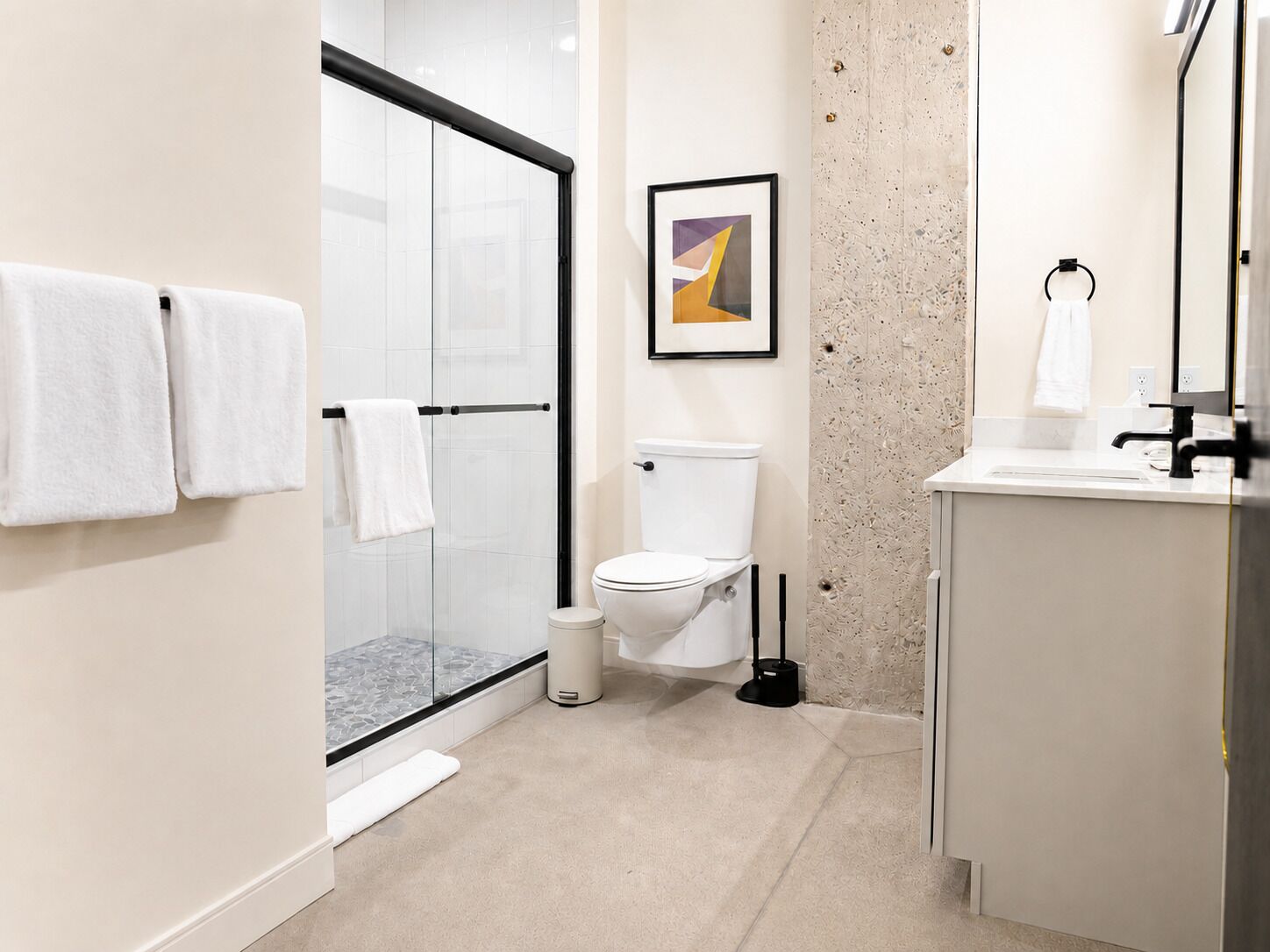 A modern bathroom with a glass shower enclosure, a toilet, a sink with a mirror, and a framed abstract painting on the wall.