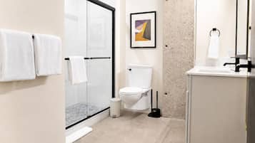 A modern bathroom with a glass shower enclosure, a toilet, a sink with a mirror, and a framed abstract painting on the wall.