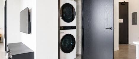 Une buanderie équipée de deux machines à laver à chargement frontal, d’une porte noire, d’un téléviseur blanc fixé au mur et d’une petite console noire.