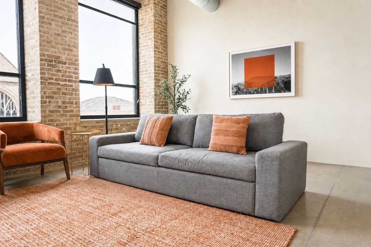 Een grijze bank met twee oranje kussens, een kleine oranje fauteuil en een groot raam met uitzicht op een gebouw.