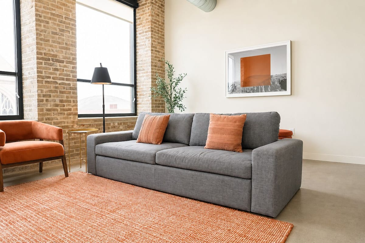 Een woonkamer met een grijze bank, oranje kussens, een klein oranje vloerkleed, een zwarte vloerlamp en een lijst met een afbeelding aan de muur.
