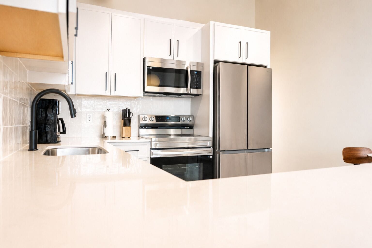 A modern kitchen with white cabinets, stainless steel appliances including a microwave, oven, and refrigerator, a black faucet, and a wooden cutting board.
