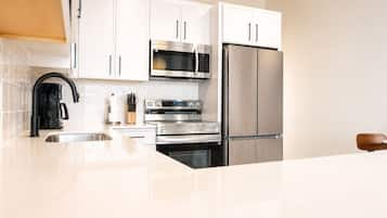 Una cocina moderna con armarios blancos, electrodomésticos de acero inoxidable, incluyendo un microondas, horno y refrigerador, un grifo negro y una tabla de cortar de madera.