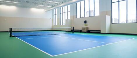 An indoor tennis court with a net, large windows, and a high ceiling.