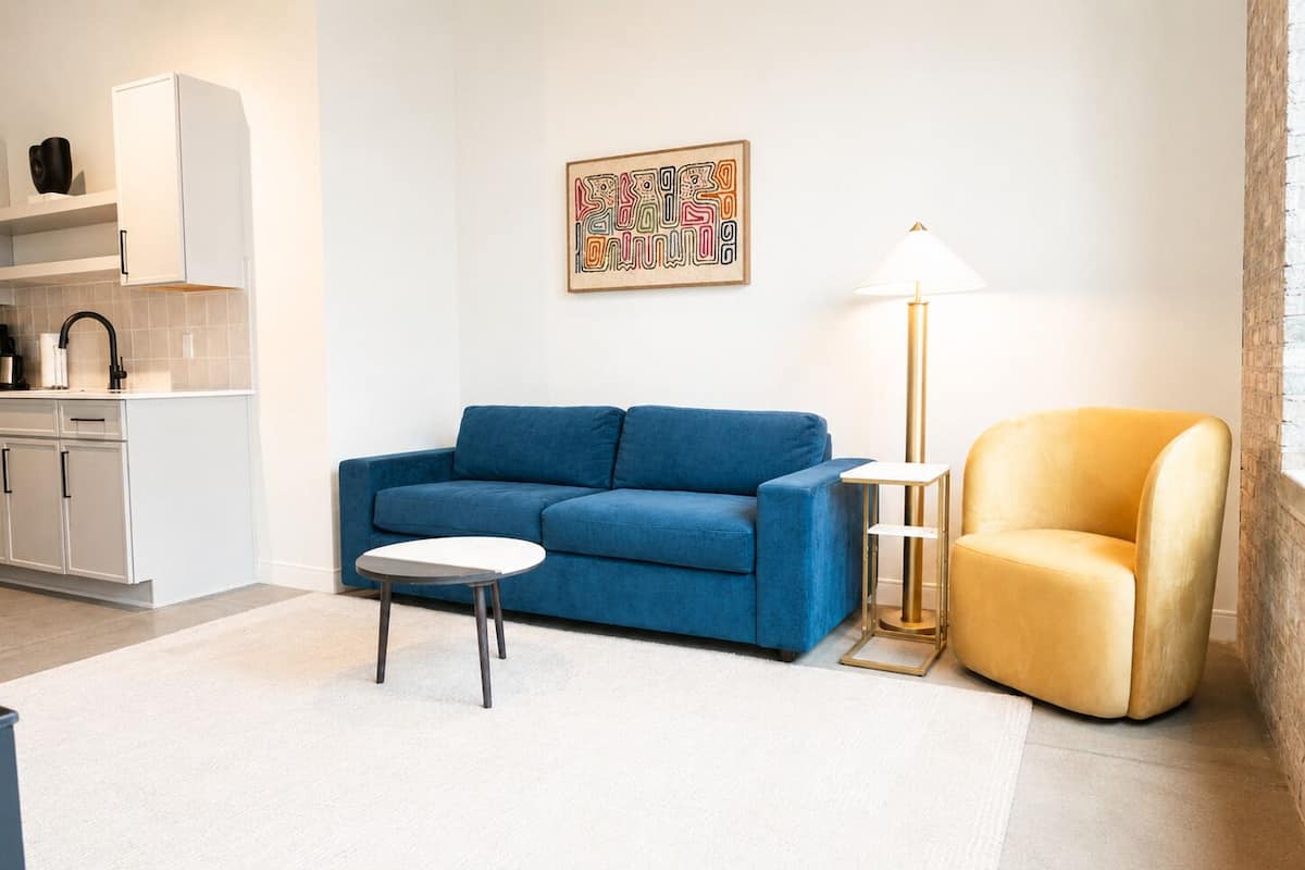 Een moderne woonkamer met een blauwe bank, een kleine witte tafel, een gele fauteuil, een vloerlamp en een keuken met witte kasten.