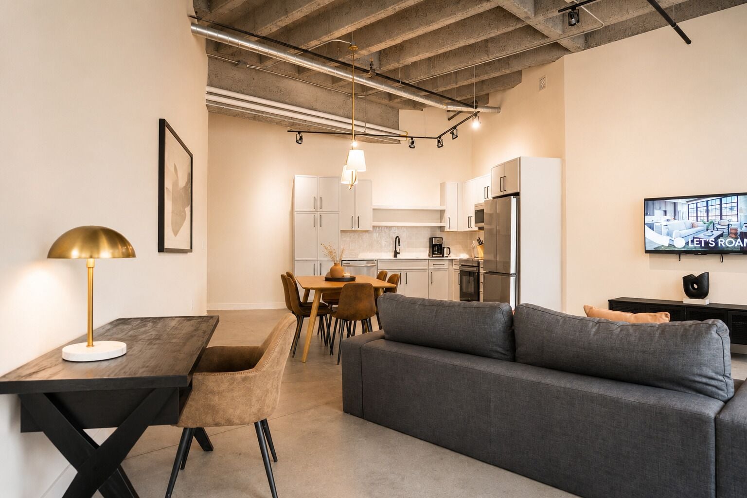 A modern kitchen with white cabinets, a dining table with chairs, a gray sofa, and a TV mounted on the wall.