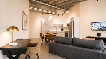A modern kitchen with white cabinets, a dining table with chairs, a gray sofa, and a TV mounted on the wall.
