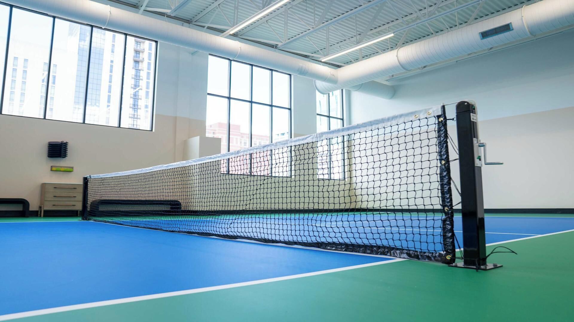 Un complexe sportif intérieur doté d’un court de tennis protégé par un filet, de grandes fenêtres et d’un plafond haut.