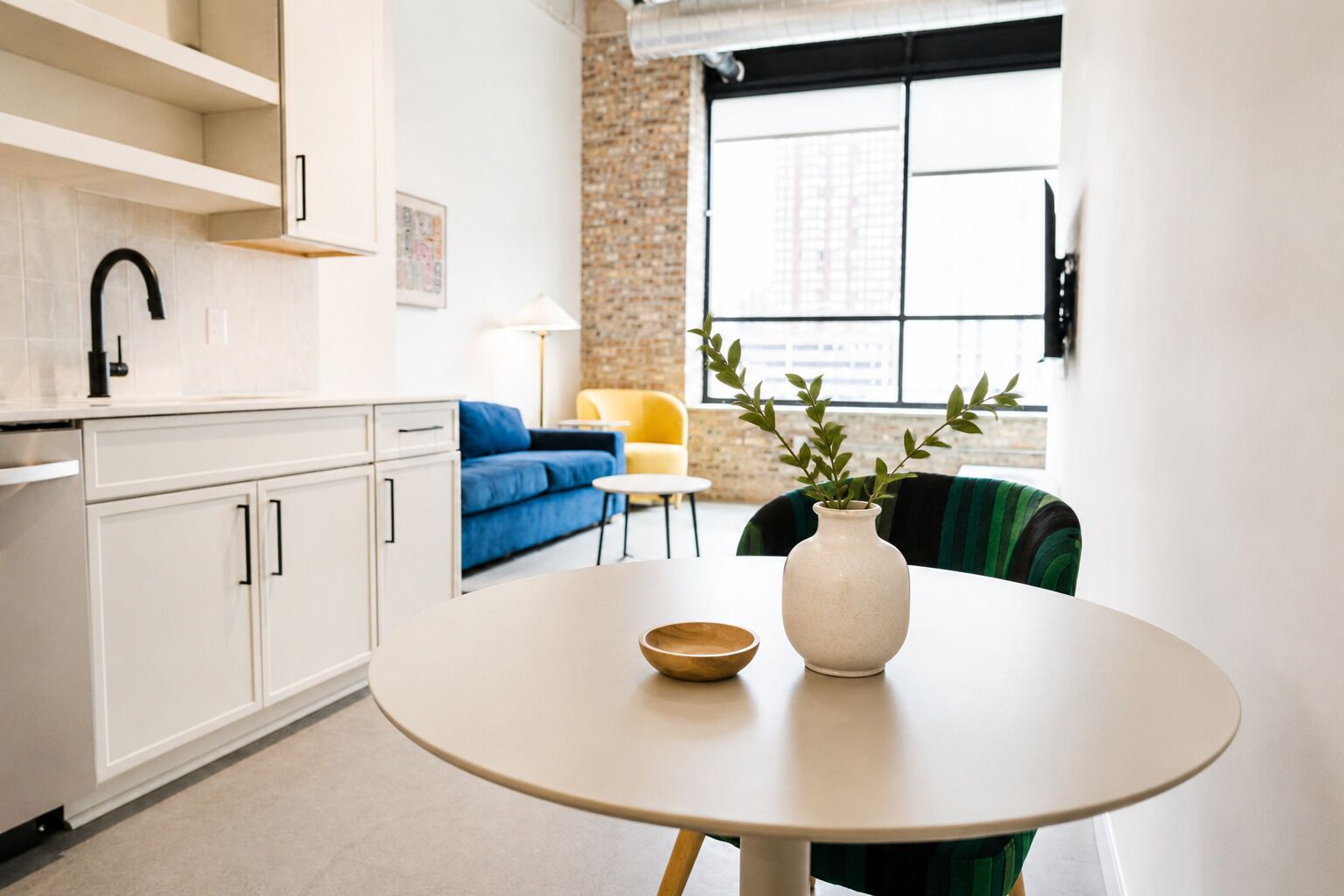 Una cocina moderna con armarios blancos, una mesa redonda con un bol y un jarrón, un sofá azul y una ventana con vistas.