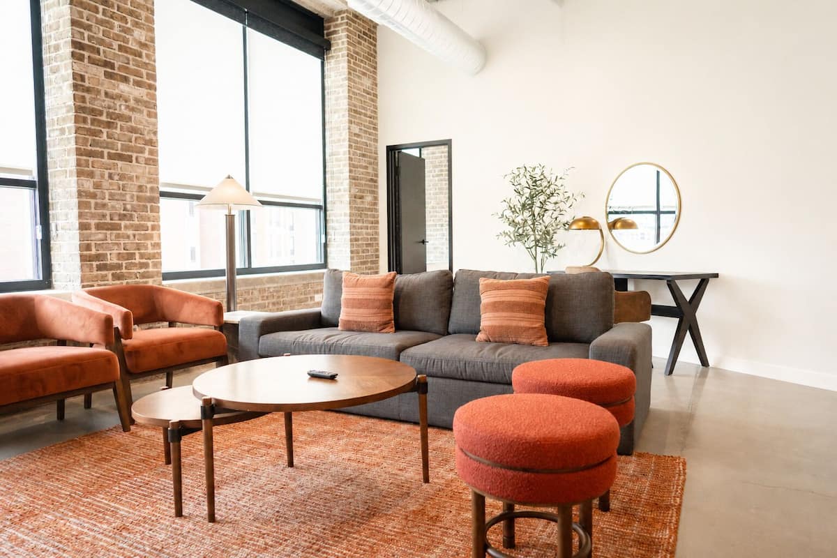 Een moderne woonkamer met een grijze bank, oranje fauteuils, een ronde salontafel en een kleine tafel met een lamp.