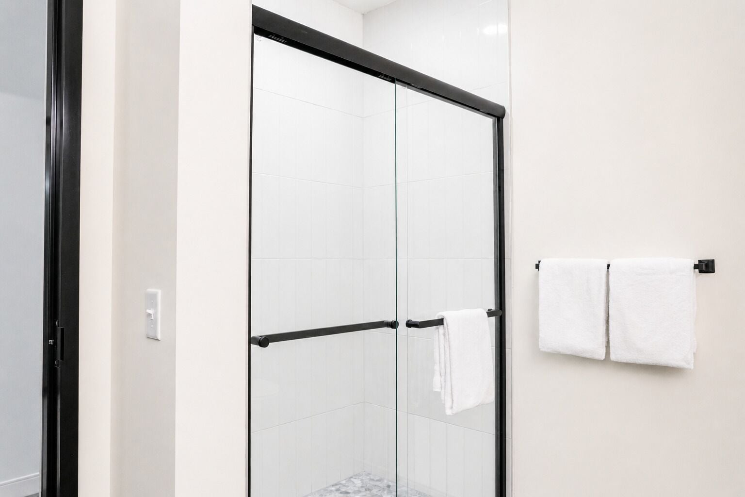 A glass-enclosed shower with a black frame, white tiled walls, and a towel rack with two white towels.