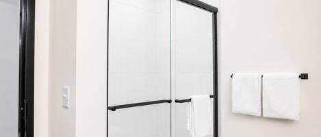 A glass-enclosed shower with a black frame, white tiled walls, and a towel rack with two white towels.