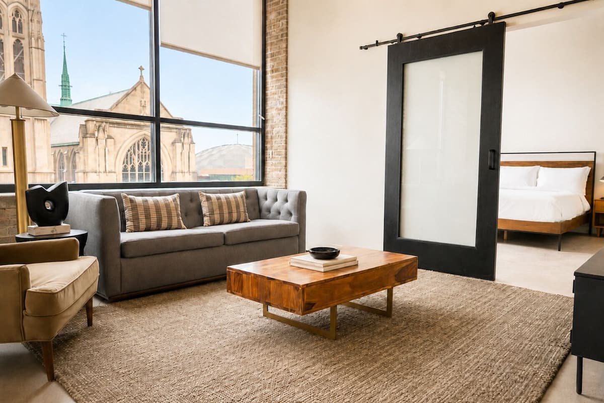 Een moderne hotelkamer met een groot raam, een grijze bank, een houten salontafel, een bed en uitzicht op een kerktoren.