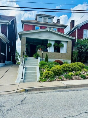 Una casa de dos pisos con un porche delantero, una escalera que conduce a la entrada y un pequeño jardín con diversas plantas.