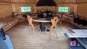 A wooden interior with a central table, chairs, a fireplace, and windows.